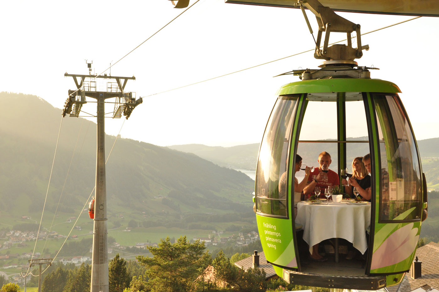 Eine geöffnete grüne Gondel an der Bergstation. In der Gondel sitzen vier Personen mit einem weiss gedeckten Tisch und stossen mit Wein an. 