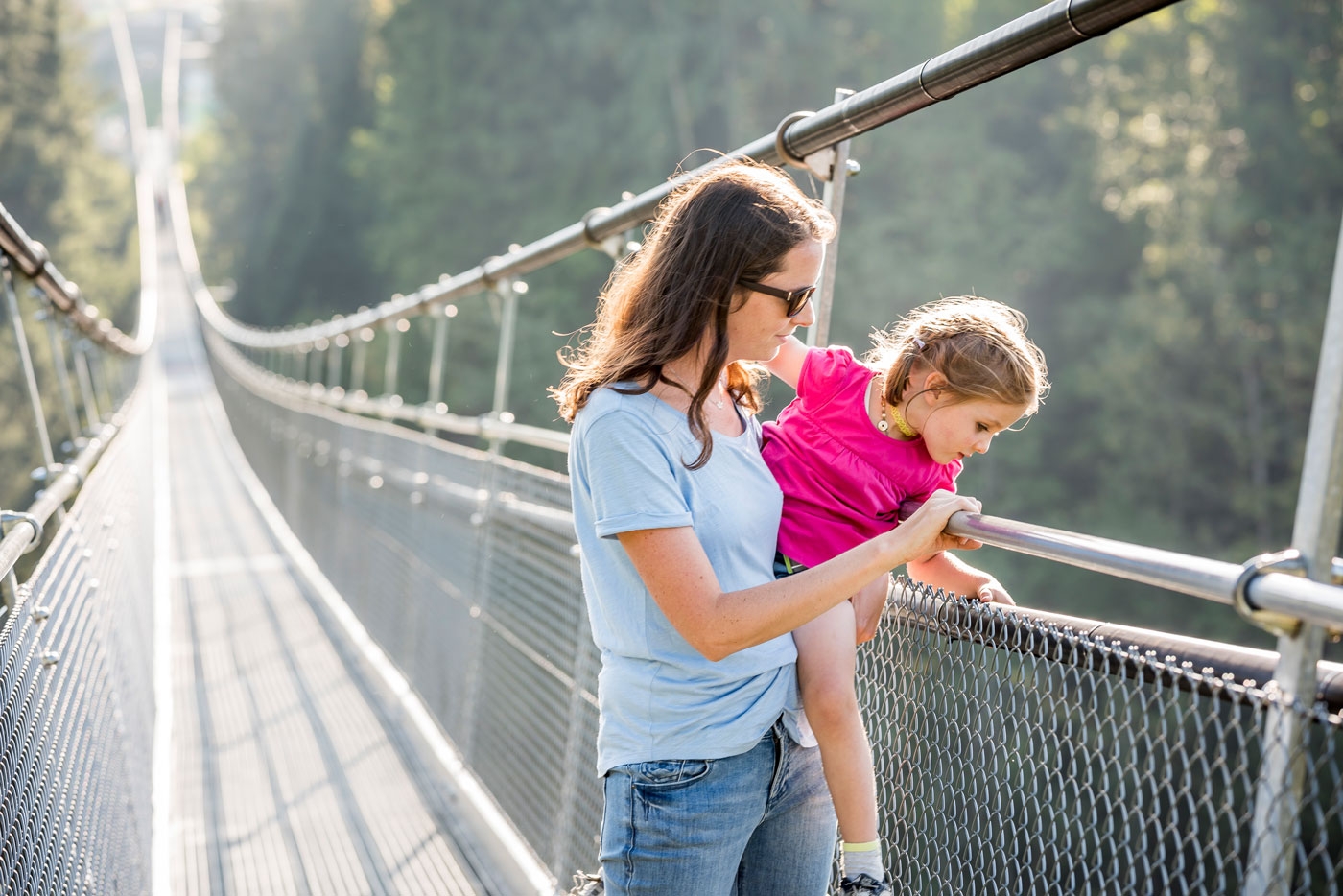 Eine Mutter hat ihre Tochter auf dem Arm und sie schauen beide von der Hängebrücke auf der sie stehen hinunter. 