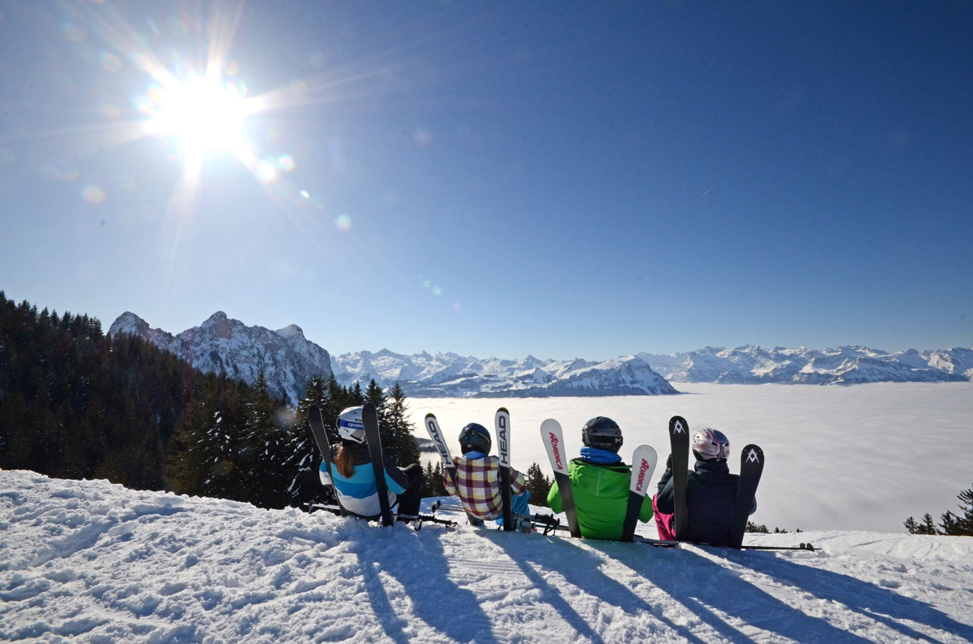 Vier Kinder sitzen mit Ihren Skiern im Schnee. Blick auf die Bergspitzen und das Nebelmeer bei Sonnenschein. 